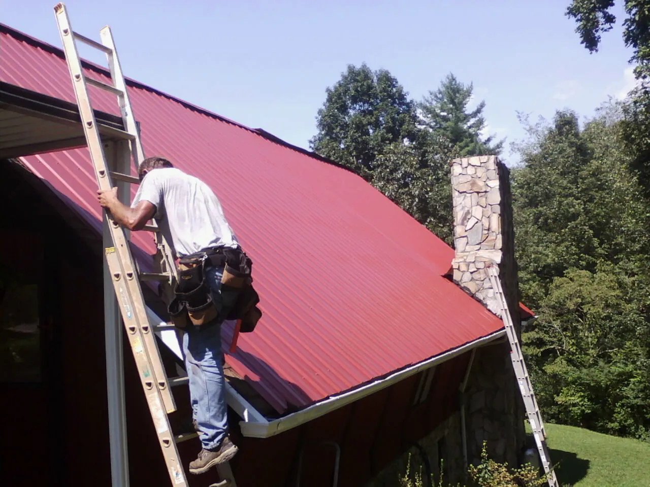 Roofer on a ladder beside a red metal roof and stone chimney — Soffit & Fascia Repair by Roofing Brothers in Stillwater